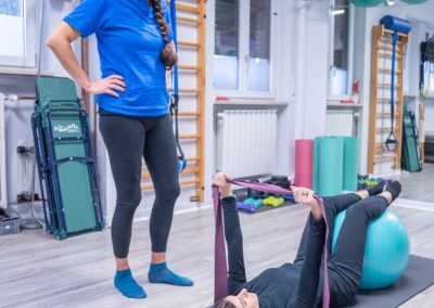 Paziente esegue esercizi riabilitativi con fitball ed elastico sotto la supervisione di un istruttore.
