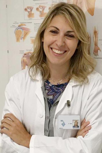Ritratto della Dott.ssa Elena Colombo, specialista in ortopedia e fisiatria, sorridente in camice bianco davanti a un poster anatomico.
