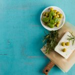 Tavola mediterranea con feta, olive verdi e basilico su fondo azzurro.
